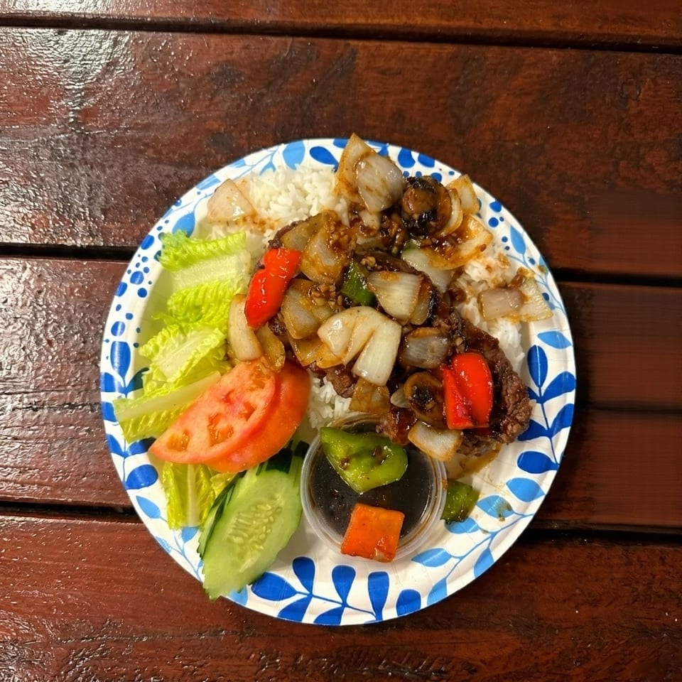 Stir-fried beef with onions and peppers, rice, lettuce, tomato, and cucumber on a paper plate.