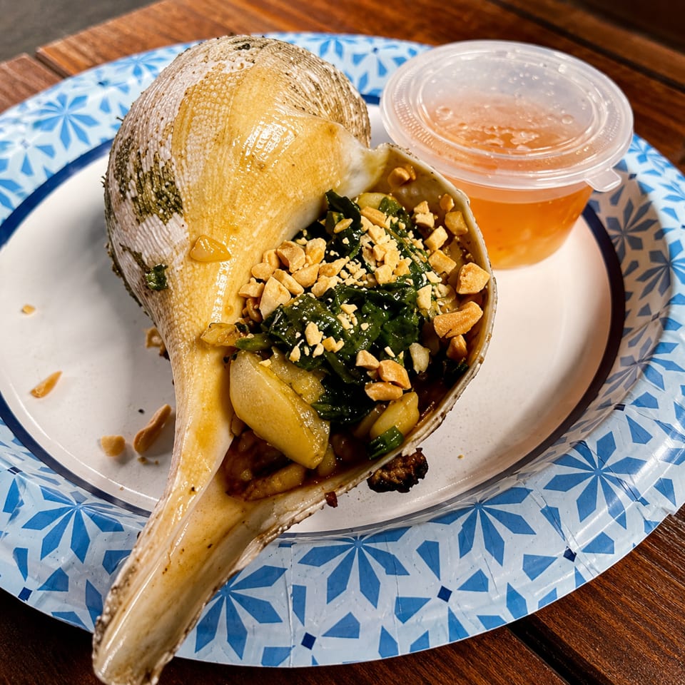 A large conch shell filled with greens, peanuts, and sauce, on a blue and white paper plate.
